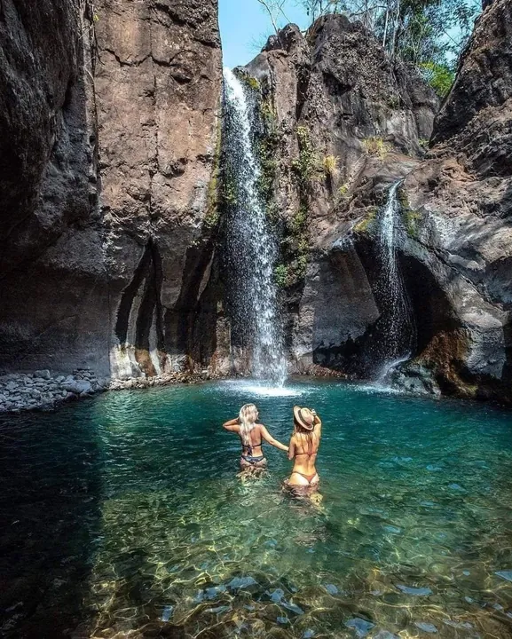 Two travelers swimming in a turquoise pool under the Tamanique waterfalls